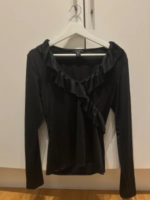 Svart blus med volang från Amisu - Säljer en svart blus från Amisu med långa ärmar och snygga volangdetaljer längs halsringningen och framtill. Blusen har en feminin och trendig look, perfekt för dig som gillar stilrena plagg med lite extra edge. Materialet är mjukt och följsamt.