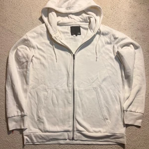 Vit hoodie med dragkedja från 157 - Vit hoodie från 157 i storlek M. Tröjan har hel dragkedja, huva med snörning och två stora fickor framtill. Tillverkad i mjuk bomullsmix som är skön att ha på sig. Perfekt för chill dagar eller när du vill ha en enkel och clean look.