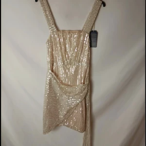 Champagne/guldfärgad klänning - Kortklänning med glittriga paljetterKlänningen har en snygg omlott-detalj med knyt till sidan. Perfekt till nyår eller något annat event. Går att dra ner och upp kjolen till en viss längd. Ingen aning vart den är ifrån tror den är köpt utomlands. 💛