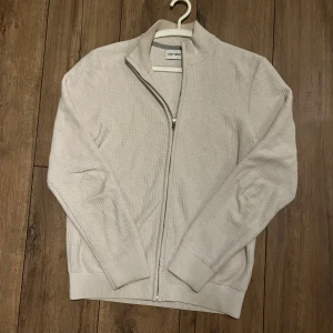 Ljusbeige vit kofta från East West - Säljer en stilren ljusbeige kofta från East West i storlek M. Koftan är stickad med ribbade muddar och har en hel dragkedja framtill. Perfekt för lager-på-lager och har en clean look med långärmad design och ståkrage. Ett perfekt val till vintern. 