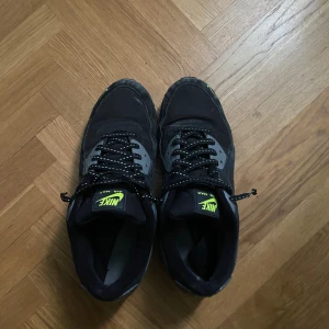 Nike Air Max svarta sneakers - Svarta Nike Air Max sneakers med grå detaljer och neongula loggor. Skorna har snörning med svarta reflex skosnören, samt en synlig Air Max-sula. Ovandelen är i syntet och mocka för en sportig look. Perfekta för dig som gillar streetstyle och klassiska Nike-vibbar.