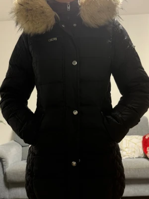 Svart lång Rockandblue dunjacka - Snygg svart lång dunjacka från Rockandblue med quiltade partier, huva med fluffig fuskpäls och flera dragkedjor. Jackan har markerad midja, långa ärmar och cool badge på ärmen. Perfekt för kalla dagar när du vill hålla stilen.
