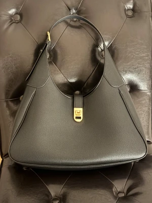 Svart axelväska med gulddetalj - Stilren svart axelväska i skinn med snygg guldfärgad låsdetalj framtill. Väskan har en mjukt rundad form och bred axelrem med guldfärgat spänne. Perfekt storlek för att få plats med allt du behöver och ger en clean vibe till din outfit.