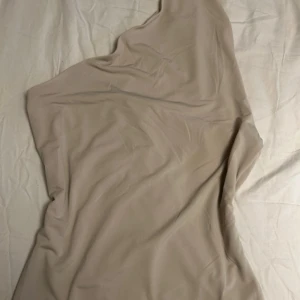 Beige oneshoulder blus/topp - Säljer en stilren beige oneshoulder klänning från H&M. Blusen har en slät och mjuk yta, med en asymmetrisk axel och enkel design. Perfekt för dig som gillar minimalistisk stil och vill ha något som sticker ut med sin unika form.