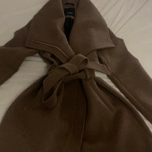 Brun kappa från Rockandblue, stl 38 - Snygg brun kappa från Rockandblue med bred krage och bälte i midjan. Jackan har en stilren design med dragkedja och stora slag. Perfekt för dig som vill ha en klassisk och trendig look under höst och vinter.