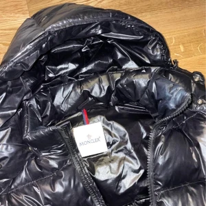 Svart Moncler pufferjacka med huva - Säljer en svart pufferjacka från Moncler med glansig finish och huva. Jackan har dragkedja framtill, två sidofickor med dragkedja och klassisk Moncler-logga på ärmen. Perfekt för kalla dagar och har en riktigt snygg streetstil.