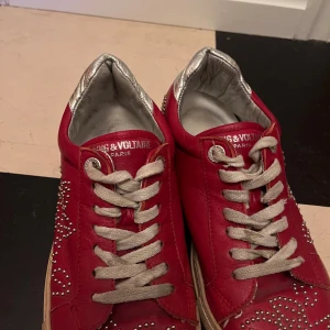 Röda Zadig & Voltaire sneakers - Snygga röda sneakers från Zadig & Voltaire med coola silverfärgade nitar i mönster på sidorna och tån. Skorna har silverfärgad häl, vita snören och beige platt sula. Tillverkade i skinn för en lyxig känsla och unik look.
