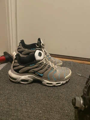 Nike Air Max Plus grå/blå sneakers - Nike Air Max Plus sneakers i grå mesh med blå och svarta detaljer. Snyggt vågigt mönster på ovansidan och vit sula med synliga luftkuddar. Skorna har snörning och rund tå, samt svart insida och vit etikett med blå swoosh på plösen.