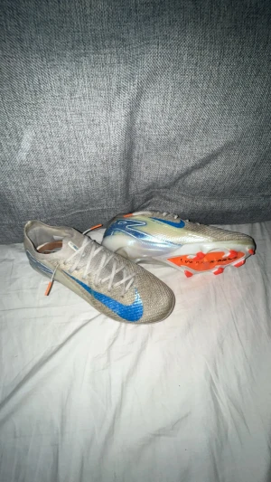 Nike Mercurial fotbollsskor blå/silver - Säljer ett par Nike Mercurial fotbollsskor i silver med blå swoosh och detaljer i orange. Skorna har snörning och platt sula med dobbar för gräsplan. Ovandelen är i syntetmaterial och designen är riktigt sportig och modern.