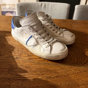 Philippe Model vita sneakers med blå detalj - Säljer ett par  vita Philippe Model sneakers i skinn med blå detalj på hälen. Skorna har snygg slitning! 