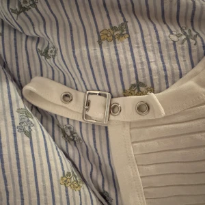 Vit ribbad linnetopp med bälte - Säljer en vit ribbad linnetopp med breda axelband som har coola bältesdetaljer i silver. 