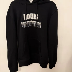 Louis Vuitton hoodie  - Svart hoodie från Louis Vuitton med stort vitt och silverfärgat tryck på bröstet. Tröjan har huva med snörning, känguruficka och lång ärm. På ryggen finns en orange patch med Louis Vuitton-logga. Perfekt för dig som gillar exklusiva streetwear-vibes.