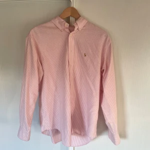 Rosa och vit randig skjorta från Polo Ralph Lauren - Snygg skjorta från Polo Ralph Lauren i rosa och vitt med smala ränder och klassisk broderad logga på bröstet. Skjortan har lång ärm, knappar framtill och en stilren krage. Perfekt för dig som gillar preppy stil och vill ha något som sticker ut.