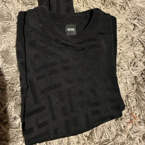 Svart stickad tröja från BOSS - Svart stickad tröja från BOSS med diskret mönster i samma färg. Rund halsringning och mjukt material som känns skönt mot huden. Perfekt för dig som gillar stilrena plagg. Storlek s men passar även M