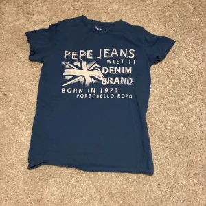 Blå t-shirt från Pepe Jeans - Snygg marinblå t-shirt från Pepe Jeans med tryck i vitt framtill. T-shirten har korta ärmar och klassisk rund hals. Materialet känns mjukt och skönt, perfekt för en chill look. Stor tryckt logga och text ger en cool street vibe.