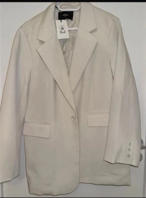 Beige kavaj från MONKI - Stilren beige kavaj från MONKI med klassisk krage och knappstängning framtill. Kavajen har två fickor med lock och tre dekorativa knappar vid ärmslutet. Perfekt för en clean och modern look.