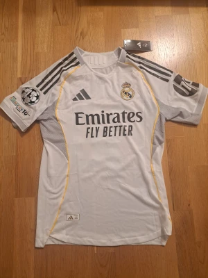 Real Madrid Mbappé 10 matchtröja - Säljer en officiell Real Madrid fotbollströja från Adidas med Mbappé och nummer 10 på ryggen. Tröjan är vit med svarta detaljer, gula linjer och Champions League-märke på ärmen. Materialet är lätt och ventilerande polyester, perfekt för match eller träning.