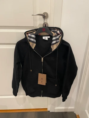 Svart hoodie från Burberry med dragkedja - Svart hoodie från Burberry med klassisk rutig huva i beige, rött och svart. Tröjan har dragkedja framtill, snörning vid huvan och två fickor. Materialet är mjuk bomull och polyester, perfekt för chill dagar. Snyggt Burberry-mönster på insidan av huvan. Passar till M och även S