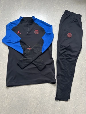 PSG x Jordan svart träningsset - Säljer en svart och blå långärmad Paris Saint-Germain träningströja från Jordan. Tröjan har röd PSG-logga på bröstet, röd Jumpman-logga och diskret Paris-tryck på ärmen. Slim fit-modell i funktionsmaterial, perfekt för träning. Pris kan diskuteras!