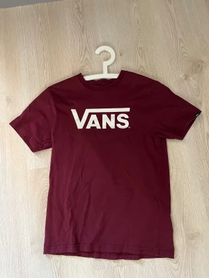 Vinröd Vans t-shirt med logga (barn) - Snygg vinröd t-shirt från Vans i storlek Large. T-shirten har klassisk passform och är tillverkad i 100% bomull. Stor vit Vans-logga tryckt på bröstet ger en clean streetstyle-look. Perfekt för dig som gillar skate och avslappnad stil.