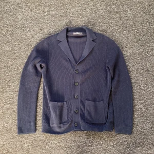 Mörkblå kofta med knappar - Snygg mörkblå kofta med ribbad struktur och klassisk krage. Den har fem svarta knappar framtill och två praktiska fickor. Perfekt för dig som gillar stilrena och enkla plagg med lite extra edge.