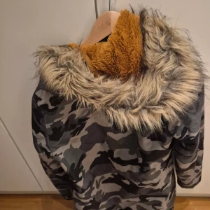 Camouflage parkasjacka med fuskpäls - Parkasjacka från Fabulous i grått och svart camouflage med mysig orange teddyfoder och stor huva med fuskpäls. Jackan har dragkedjor framtill, stora fickor och en cool streetstil. Perfekt.
