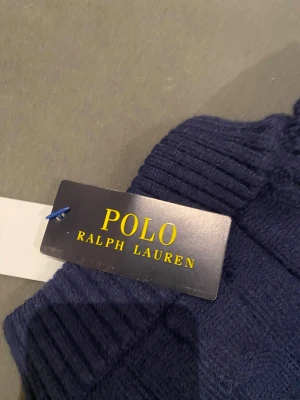 Marinblå stickad tröja Polo Ralph Lauren - Snygg marinblå stickad tröja från Polo Ralph Lauren med kabelstickat mönster och röd logga på bröstet. Tröjan har lång ärm, ribbade muddar och en half zip vid halsen för extra stilpoäng. Perfekt för dig som gillar klassisk och clean look.