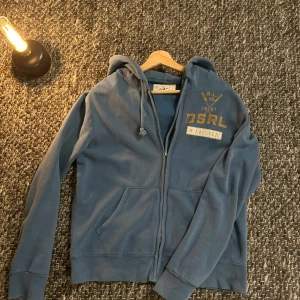 Denim and supply zip hoodie - Snygg blå hoodie från Denim & Supply Ralph Lauren med gul vintage-tryck både fram och bak. Tröjan har huva med snörning, dragkedja och två fickor framtill. Tillverkad i mjuk bomull, perfekt för chill dagar. Storlek M.