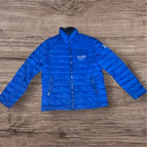 Blå dunjacka från Salming XL - Blå dunjacka från Salming i storlek XL med quiltad design och dragkedja framtill. Jackan har två sidofickor med dragkedja och en bröstficka med tryckknappar. Materialet är syntet och jackan har hög krage. Perfekt för dig som gillar sportig stil.