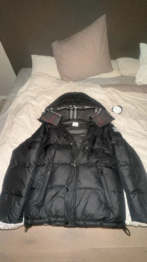 Svart pufferjacka från Burberry - Svart pufferjacka från Burberry med huva och klassiskt rutigt foder i kragen. Jackan har dragkedja och knappar framtill, samt två fickor med dragkedja. På vänster ärm finns Burberry London England-tryck. Perfekt för kalla dagar.