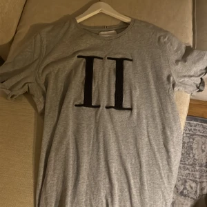 Grå t-shirt från Les Deux med tryck - Snygg grå t-shirt från Les Deux med stort svart II-tryck på bröstet. Klassisk rund hals och korta ärmar. Mjuk och skön bomullskvalitet som passar perfekt till jeans eller shorts. Enkel men stilren design med en modern vibe.