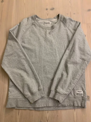 Grå sweatshirt från Resteröds XL - En stilren grå sweatshirt från Resteröds i storlek XL men passar L. Tröjan har rund hals, långa ärmar och en diskret logotyp-patch vid nederkanten. Tillverkad i mjuk bomullsmix som är skön mot huden och passar perfekt för chill dagar. Enkel och clean design utan mönster.