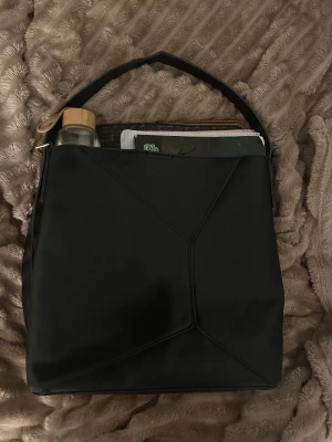 Svart axelväska i skinn - Stilren svart axelväska med geometriska sömmar och bred axelrem. Väskan är rymlig och har plats för laptop, anteckningsblock och vattenflaska. Materialet är slätt skinn och designen är minimalistisk med en modern touch.