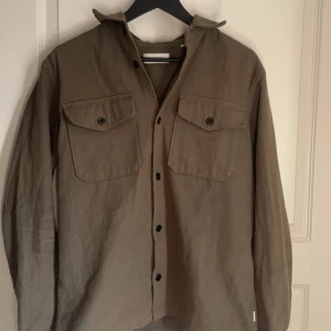 Brun långärmad skjorta med knappar - Snygg grön långärmad skjorta med knappar och två bröstfickor. Perfekt för en stilren look. Skjortan har en klassisk krage och e riktigt snygg.