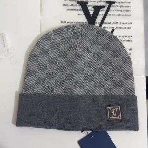 Grå rutig L och V mössa - Snygg grå mössa från L och V med rutigt mönster och uppvikt kant. L och V-logga i grått och beige framtill. Perfekt accessoar för att hålla dig varm och stilren under kyliga dagar.