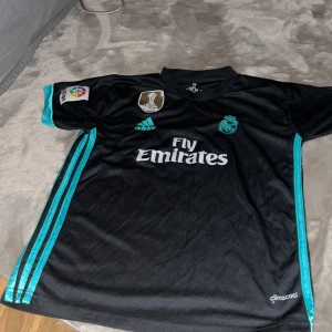 Real Madrid Ronaldo 7 svart matchtröja - Svart Real Madrid fotbollströja från Adidas med Ronaldo 7-tryck på ryggen. Tröjan har turkosa detaljer och ränder längs sidorna, samt Fly Emirates-logga på bröstet. ClimaCool-material som andas och korta ärmar. Perfekt för dig som älskar fotboll och Cristiano Ronaldo.