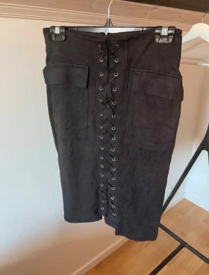 Svart kjol med snörning - Snygg svart kjol med cool snörning framtill. Kjolen har en tight passform och är gjord i ett mjukt material som liknar mocka och stretch. Perfekt för dig som gillar detaljer och vill sticka ut lite extra.