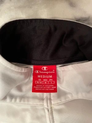 Vit vindjacka från Champion M - Snygg vit vindjacka från Champion i storlek medium. Jackan har svarta detaljer längs ärmarna med Champion-loggor, hög krage och dragkedja framtill. Tillverkad i polyester och har två fickor på sidorna. Perfekt för en sportig och clean look.