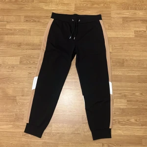 Svarta och beige mjukisbyxor från BOSS - Snygga mjukisbyxor från BOSS i svart med beige paneler längs sidorna och vita diagonala detaljer på benen. Byxorna har snörning i midjan och muddar vid bensluten för en skön passform. Perfekta för chill eller träning. KÖP HELA DRESSEN OCH FÅ DE FÖR 1500kr