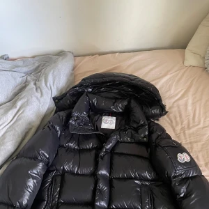 Svart pufferjacka från Moncler - Säljer en svart, glansig pufferjacka från Moncler med klassisk logga på ärmen och innerfoder med serietidningsmönster. Jackan har hög krage, dragkedja och stora vadderade partier som ger en riktigt fet vinterlook. Perfekt för dig som vill ha både stil och värme. Både nfc och qr Scan finns. Perfekt inför hösten och vintern.