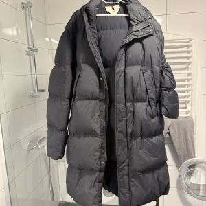 Svart lång dunjacka från Arket - Säljer en svart, lång dunjacka från Arket med fyllning från Re:Down. Jackan har huva, dragkedja och knappar framtill samt stora, quiltade sektioner. Perfekt för kalla vinterdagar och har en clean, oversized look. Obs finns två små hål som du ser på sista bilden. 