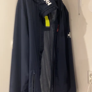 Mörkblå Helly Hansen vindjacka - Snygg mörkblå vindjacka från Helly Hansen med Helly Tech-tryck på ärmen och huva. Jackan har meshfoder, dragkedja framtill och praktiska fickor. Perfekt för blåsiga dagar och har en sportig vibe med stilrena detaljer. Storlek M