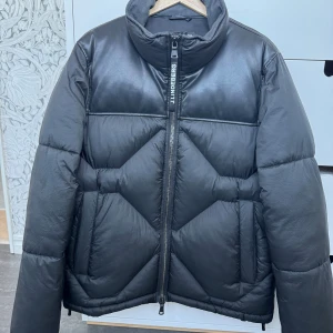 Svart pufferjacka från J.Lindeberg - Snygg svart pufferjacka från J.Lindeberg med cool quiltad design och hög krage. Jackan har detaljer i skinn på axlar och bröst, samt en rejäl dragkedja framtill med logotyp. Perfekt för dig som vill ha en stilren, modern och varm jacka.