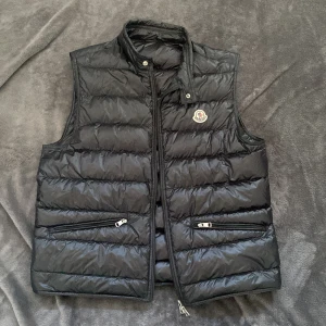 Svart Moncler dunväst med dragkedja - Svart dunväst från Moncler, modell GUI GILET. Västen har quiltad design, hög krage med knapp, två fickor med dragkedja och Moncler-logga på bröstet. Materialet är glansig polyester och dunfyllning. Perfekt för lager-på-lager och snygg till jeans eller hoodie.