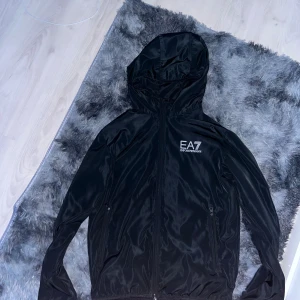 Svart EA7 vindjacka med huva - Snygg svart vindjacka från EA7 Emporio Armani med huva och dragkedja framtill. Jackan har två sidofickor med dragkedja och en stilren logga på bröstet. Materialet är lätt och glansigt, perfekt för en sportig look.