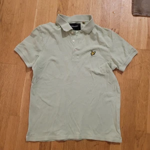 Ljusgrön piké från Lyle & Scott - Fräsch ljusgrön pikétröja från Lyle & Scott med klassisk krage och tre knappar framtill. På bröstet sitter den ikoniska broderade gula fågeln. Tröjan är kortärmad och gjord i mjuk bomull som är skön mot huden. Perfekt för en chill och stilren look.
