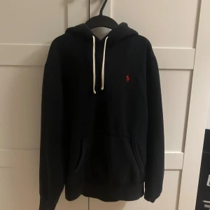 Svart hoodie från Polo Ralph Lauren - Svart hoodie från Polo Ralph Lauren med vit snörning och röd broderad logga på bröstet. Tröjan är i använt skick och smått blekt över tiden men annars i bra skick därav priset