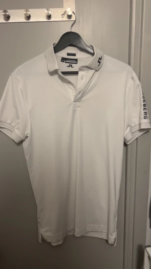 Vit J.Lindeberg piké med tryck - Fräsch vit pikétröja från J.Lindeberg med svart logotyp på kragen och ryggen samt tryck på ena ärmen. Klassisk krage, korta ärmar och knappar framtill. Materialet är mjukt och lätt polyester som andas bra. Perfekt för dig som gillar stilren sportig look.