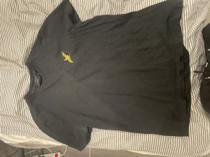 Svart t-shirt från All Saints - Svart t-shirt från All Saints med korta ärmar och rund hals. På bröstet finns ett broderat gult fågelmotiv som ger en cool detalj. T-shirten är gjord i mjuk bomull och har en klassisk, normal passform. Perfekt för dig som gillar stilrena plagg med en twist.