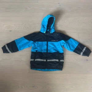 Blå och svart fodrad regnjacka men även mörkblåa regnbyxor  regnbyxor från Tenson - Snygg orginal blå och svart fodrad regnjacka men även mörkblåa regnbyxor från Tenson med huva och reflexdetaljer. Jackan har dragkedja framtill, två fickor med dragkedja och elastiska muddar. Perfekt för regniga dagar. Storlek 122/128 i både byxor och jacka det är ett sett.